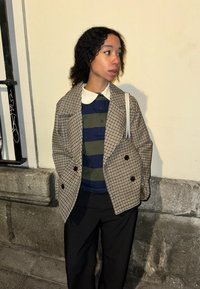 Veste à double boutonnage à carreaux beige et marron, portée sur un polo rayé vert et marine, assortie avec un pantalon noir et un sac à main blanc.