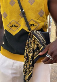 Μαύρο και κίτρινο πλεχτό γιλέκο σε σχέδιο argyle, συνδυασμένο με μια μαύρη τσάντα που διαθέτει χρυσές λεπτομέρειες και μια μαύρη γούνα. Ασημένιο δαχτυλίδι στο χέρι.