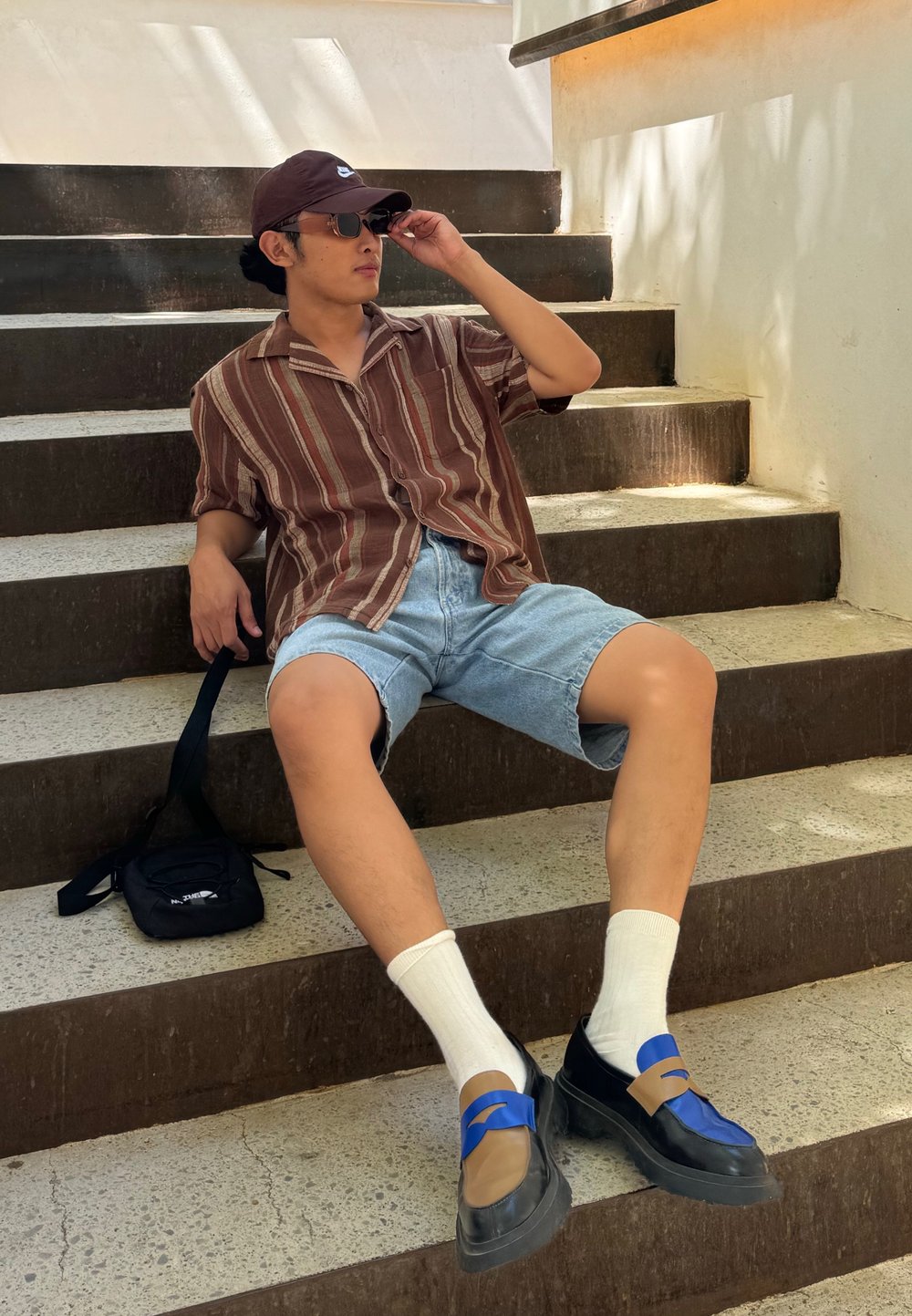 Camicetta a maniche corte a righe marrone e beige, pantaloni corti di jeans, calzini bianchi e scarpe nere con dettagli blu e marroni, seduto su gradini di pietra.