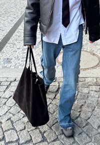 Bolsa tote de camurça preta com duas alças longas, segurada por uma pessoa vestindo uma jaqueta de couro preta, camisa branca e calças de ganga. Pavimento de pedras.