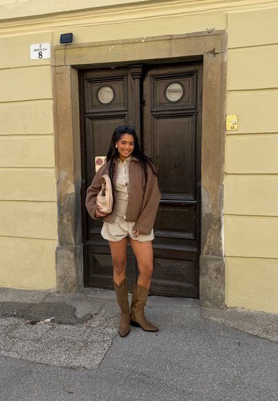 Mujer joven con chaqueta marrón, vestido de cuadros y botas, sonriendo y sosteniendo un bolso beige frente a una puerta de madera oscura en una calle de la ciudad.