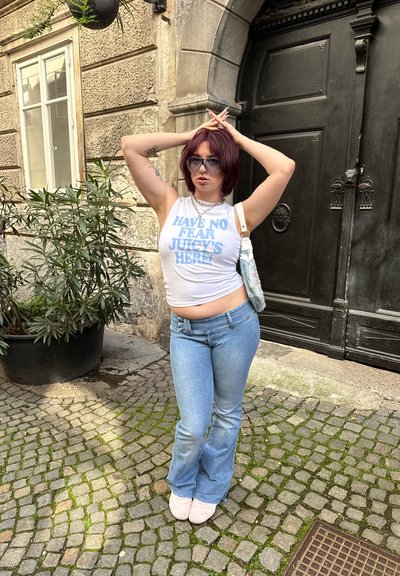 Top blanco sin mangas con texto azul, jeans claros acampanados y zapatos rosa suave. Gafas de sol negras y un bolso blanco de hombro. El texto dice "¡NO TENGAS MIEDO, JUICY ESTÁ AQUÍ!"