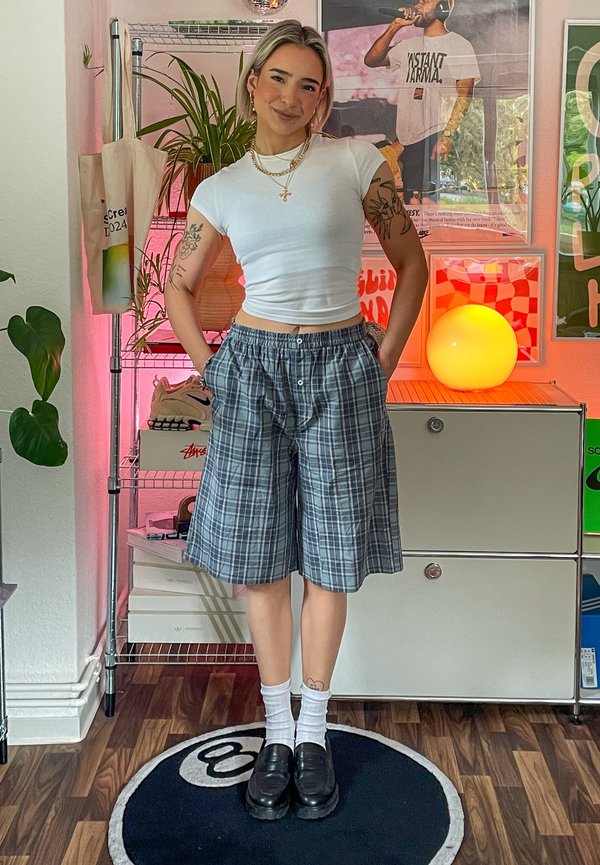 Grijze geruite shorts met een elastische tailleband, gecombineerd met een getailleerde witte blouse. Zwarte instapper, witte sokken en een casual binnenomgeving.