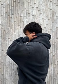Personne aux cheveux foncés portant un sweat à capuche noir, ajustant la capuche, debout devant un mur en briques grises claires texturé.