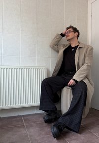 Homme portant des lunettes, manteau beige, chemise noire et pantalon large à carreaux, assis à l'intérieur sur un ottoman rond, main posée sur la tête.