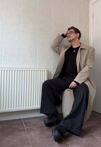 Hombre con gafas, abrigo beige, camisa negra y pantalones anchos de cuadros sentado en el interior sobre un otomán redondo, apoyando la mano en la cabeza.