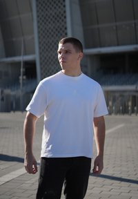 Bílá bavlněná tričko s malým logem na hrudi, spárované s černými roztrhanými džíny. Venkovní prostředí s moderní architektonickou kulisou.