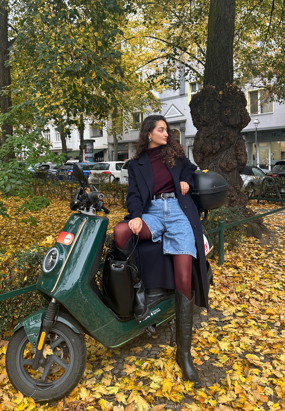 Zelen električni skuter s okroglo sprednjo lučjo in oranžnimi poudarki, parkiran na jesenskih listih. Oseba v dolgem plašču in čevljih do kolen sedi ob njem.