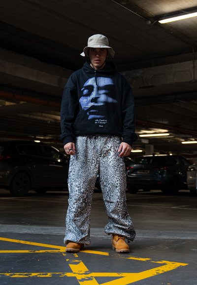 Sudadera negra con un gráfico de rostro azul y texto, combinada con pantalones de leopardo oversized y botas de trabajo color beige. Un sombrero de cubo blanco completa el look.