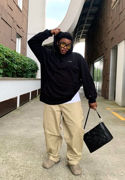 Sudadera con capucha negra, pantalones sueltos beige, zapatillas beige y un bolso tejido negro texturizado. El fondo presenta vegetación y arquitectura moderna.