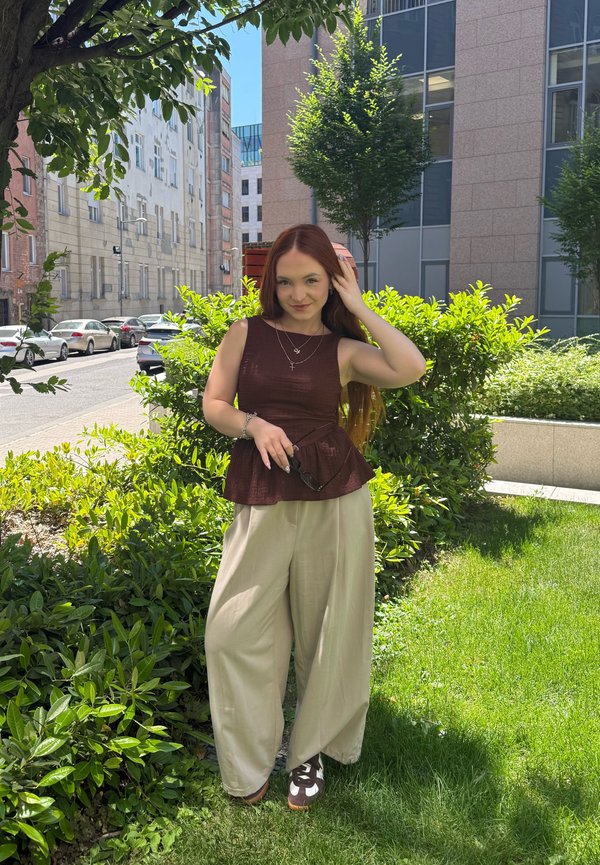 Jeune femme aux longs cheveux roux debout sur l'herbe près de buissons dans un cadre urbain, portant un haut sans manches foncé, un pantalon large beige et tenant des lunettes de soleil.