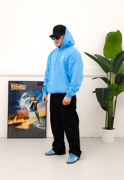 Sudadera azul claro con bolsillo frontal, pantalones holgados negros y zapatillas azules con rayas blancas. Lleva puesto una gorra negra y gafas de sol.