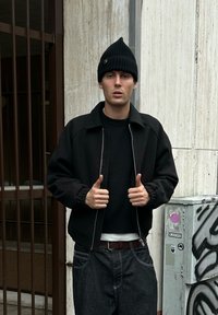 Jeune homme portant un bonnet noir, une veste noire et un jean foncé, debout avec un pouce levé dans un cadre urbain près de bars et d'un boîtier utilitaire couvert de graffitis.