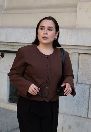 Chaqueta marrón abotonada con mangas de campana, combinada con pantalones negros. La persona sostiene gafas de sol y un pequeño bolso.