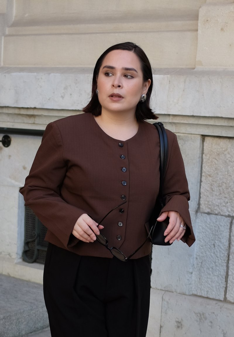Braune Knopfjacke mit Glockenärmeln, kombiniert mit schwarzen Hosen. Die Person hält eine Sonnenbrille und eine kleine Handtasche.
