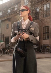 Blazer cinza oversized com um arnês, camisa branca e calças de ganga escuras. Os acessórios incluem óculos de sol pretos e uma pequena bolsa preta.