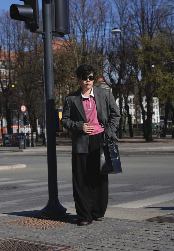 Person med solbriller, grå blazer, rosa skjorte og svarte bukser står ved et trafikklys i en bygate.