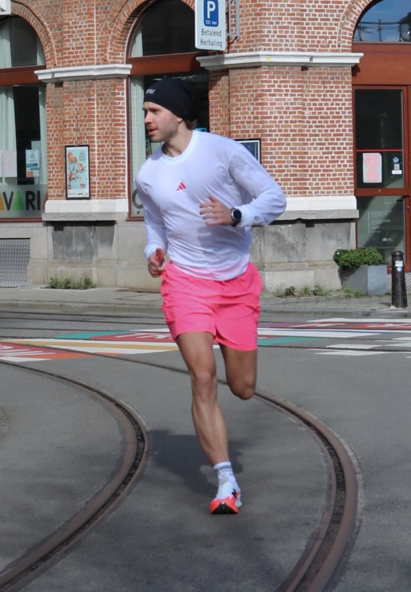 Mann in weißem Langarmhemd und rosa Shorts läuft auf Straßenbahnschienen neben einem Backsteingebäude mit Parkzeichen darüber.