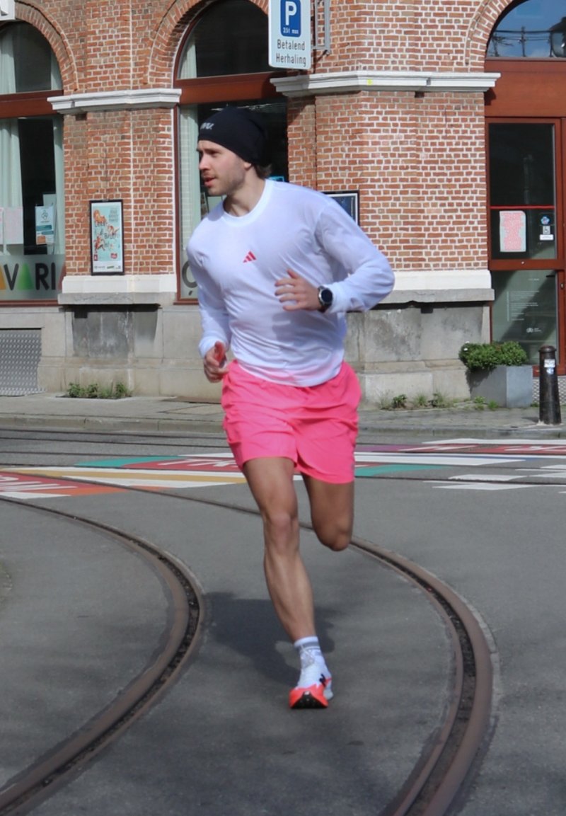 Muž v bielej košeli s dlhým rukávom a ružových kraťasoch beží po koľajniciach električky pri tehlovej budove so značením parkoviska nad hlavou.