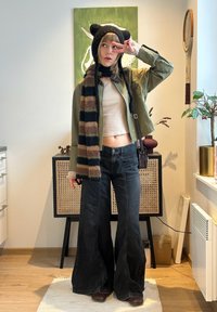 Jeune personne portant un chapeau à oreilles de chat, écharpe rayée, top beige court, veste verte, jean noir large, posant avec un signe de paix en intérieur.