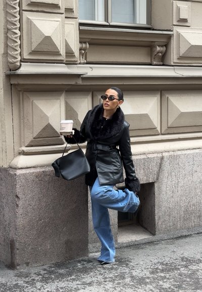 Mujer con chaqueta de cuero con cuello de piel negra, vaqueros azules y gafas de sol apoyada contra un edificio de piedra decorativa, sosteniendo un café y un bolso negro.
