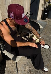 Burgundska kapa s prednjim vezom, crna majica bez rukava, tetovaže na rukama u uzorcima, crne široke hlače i smeđe-bijele cipele. Kvadratni stol u vanjskom okruženju.