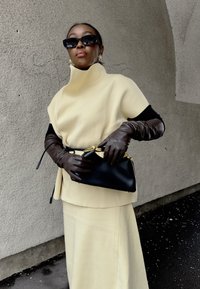 Femme portant un haut crème à col montant et une jupe, des gants longs marron foncé, une ceinture noire et un sac à main, avec des lunettes de soleil noires, devant un mur texturé.