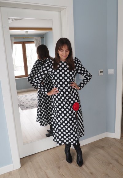 Mujer con vestido de lunares blancos y negros, con detalle de rosa roja, de pie con las manos en las caderas frente a un espejo de cuerpo entero.