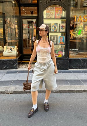 Top de tirantes blanco de encaje con acentos rosas, pantalones cortos de sudadera grises, mocasines marrones con detalle de cadena y un bolso de hombro marrón. Entorno urbano al aire libre.