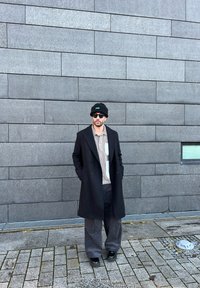 Cappotto nero sopra una camicia a fantasia con taschino, pantaloni larghi scuri e un berretto nero, indossato con occhiali da sole, contro un muro di pietra.