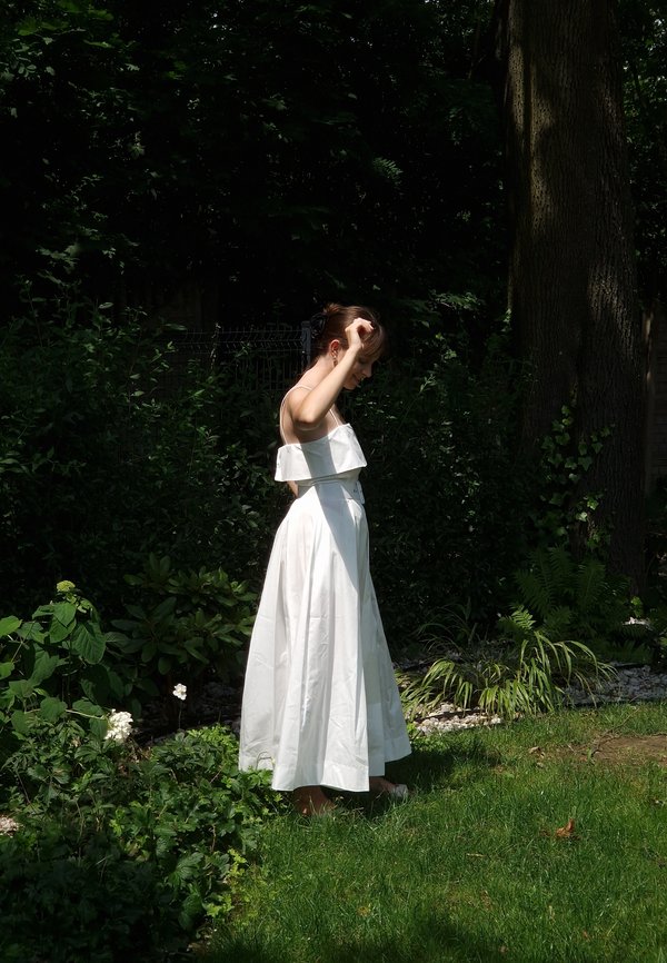 Hvid, knælængde kjole med en tætsiddende overdel og off-the-shoulder flæset halsudskæring. Letvægtsstof, flagrende skørt, barefods på græs.
