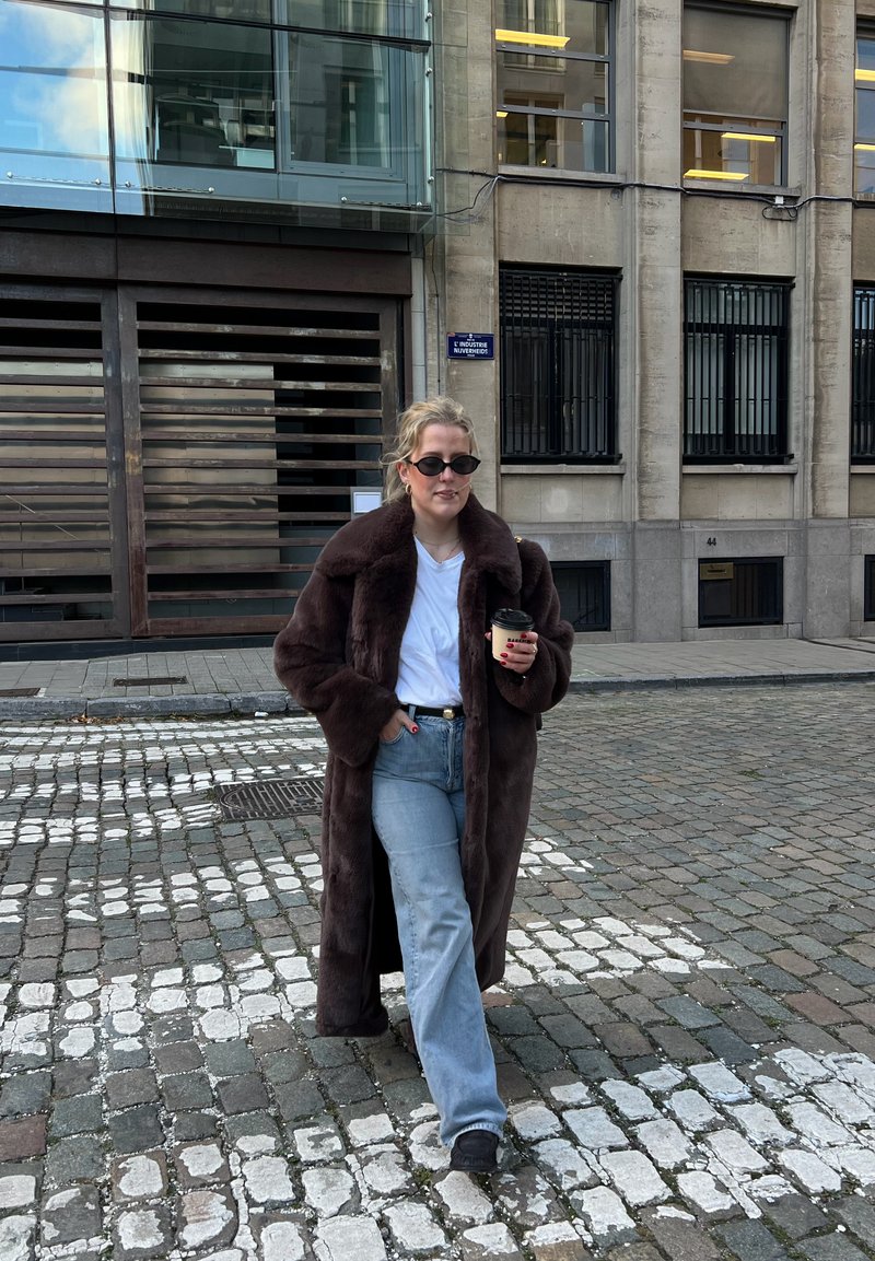 Femme portant un manteau en fourrure brun foncé, une chemise blanche et des lunettes de soleil, marchant sur une rue pavée en tenant une tasse de café près d'un bâtiment beige.