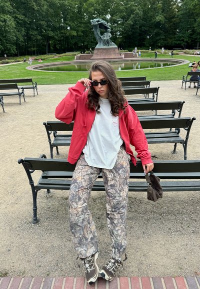 Sudadera roja con cremallera, camiseta blanca, pantalones de camuflaje y botas de senderismo marrones; gafas de sol negras y bolsa marrón en la mano; entorno de parque.