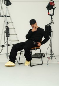 Maglietta nera a maniche lunghe, pantaloni neri e sneakers gialli. Seduto su una sedia, con un'impostazione di illuminazione e una scala sullo sfondo.