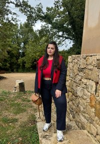 Rossa sopra con un blazer blu scuro, jeans scuri e sneakers bianche. Tiene una piccola borsa marrone. In piedi accanto a un muro di pietra, circondata da verde.