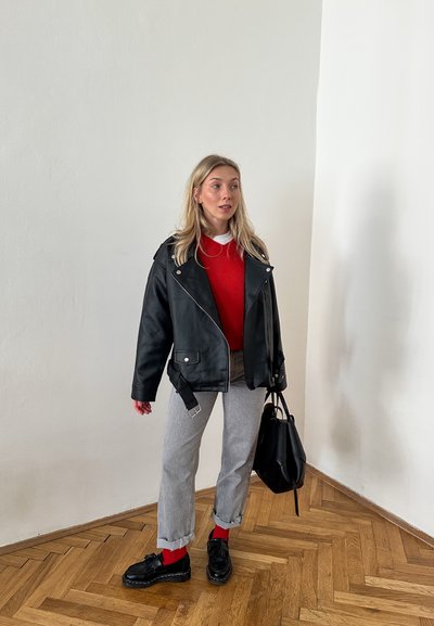 Chaqueta de cuero negra de motorista sobre un suéter rojo, pantalones grises con los puños remangados y zapatos negros. Se lleva un bolso negro, frente a una pared lisa.