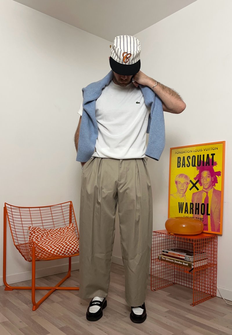 Homme portant une casquette à fines rayures, un t-shirt blanc, un pantalon large beige et un pull bleu drapé sur les épaules, debout à côté d'une chaise en fil orange et d'une affiche colorée de Basquiat.