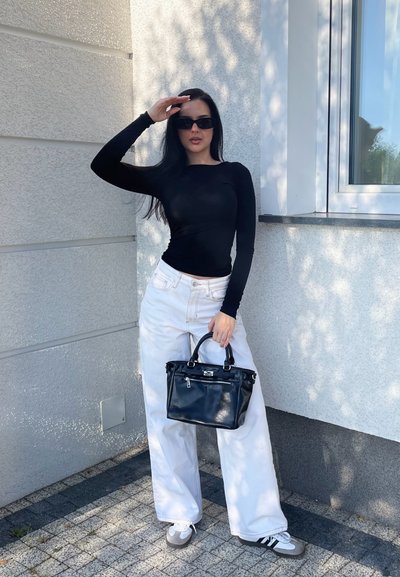 Top negro de manga larga, jeans blancos de pierna ancha y bolso negro. El fondo muestra una pared gris claro y una ventana con sombras.