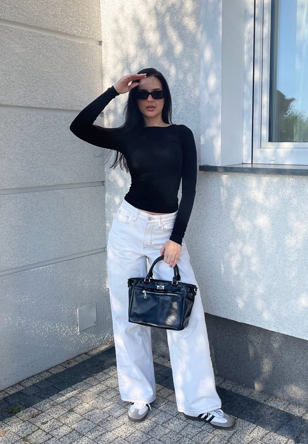 Schwarzes Langarmoberteil, weiße weit geschnittene Jeans und schwarze Handtasche. Der Hintergrund zeigt eine hellgraue Wand und ein Fenster mit Schatten.