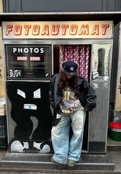 Persona con chaqueta de cuero negra y gorra frente a una cabina fotográfica con el letrero "FOTOAUTOMAT" y una cortina con patrón.