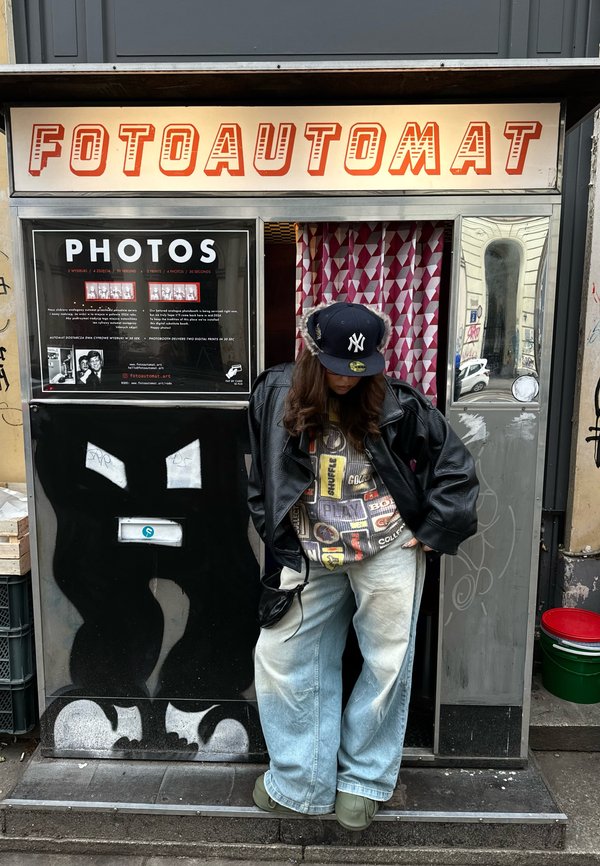 Osoba w czarnej skórzanej kurtce i czapce stoi przed budką fotograficzną z napisem „FOTOAUTOMAT” i wzorzystą zasłoną.