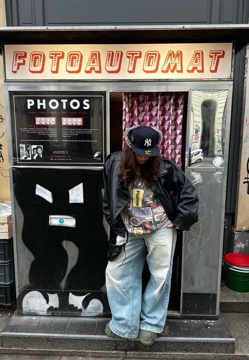 Person in einer schwarzen Lederjacke und Mütze steht vor einem Fotokabinenautomaten mit der Aufschrift "FOTOAUTOMAT" und einem gemusterten Vorhang.