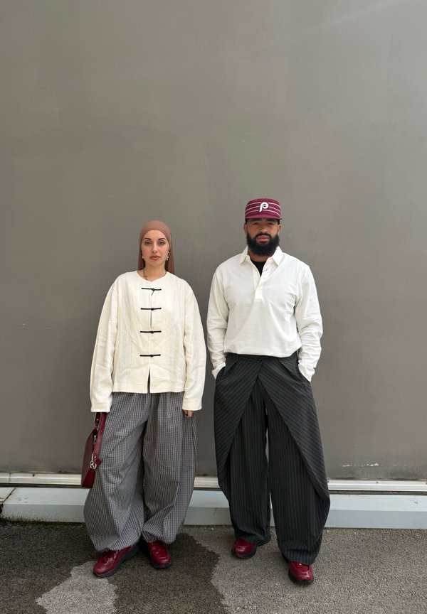 Deux personnes se tiennent contre un mur gris, portant des hauts blancs, des pantalons larges à carreaux, des chaussures rouges, l'une avec un foulard et l'autre avec une casquette rouge.