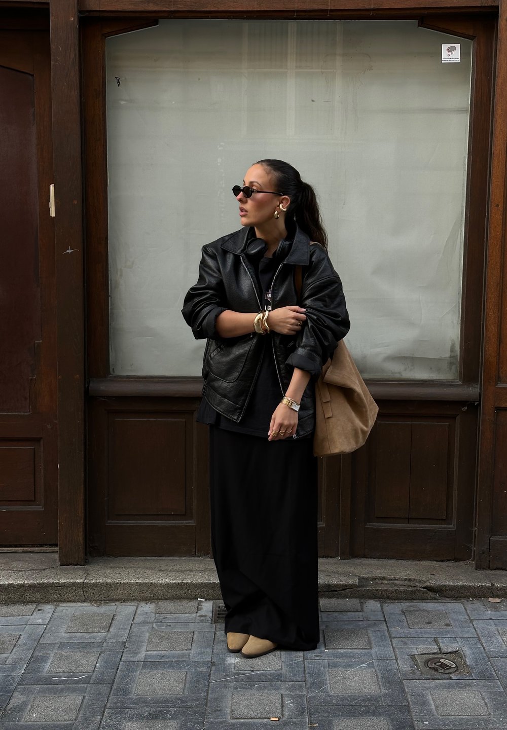 Μαύρο δερμάτινο oversized μπουφάν πάνω από ένα μαύρο φόρεμα, με αξεσουάρ χρυσά βραχιόλια, μια καφέ σουέτ τσάντα και στρογγυλά γυαλιά ηλίου, σε φόντο ξύλου.
