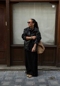 Μαύρο δερμάτινο oversized μπουφάν πάνω από ένα μαύρο φόρεμα, με αξεσουάρ χρυσά βραχιόλια, μια καφέ σουέτ τσάντα και στρογγυλά γυαλιά ηλίου, σε φόντο ξύλου.