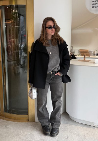 Mujer con abrigo negro, suéter gris, jeans oscuros, botas negras y gafas de sol, sosteniendo un pequeño bolso plateado junto a un mostrador blanco.
