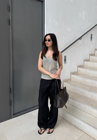 Top sin mangas de color gris claro, pantalones de pierna ancha negros, bolso oscuro y gafas de sol; de pie cerca de una escalera y una puerta gris.