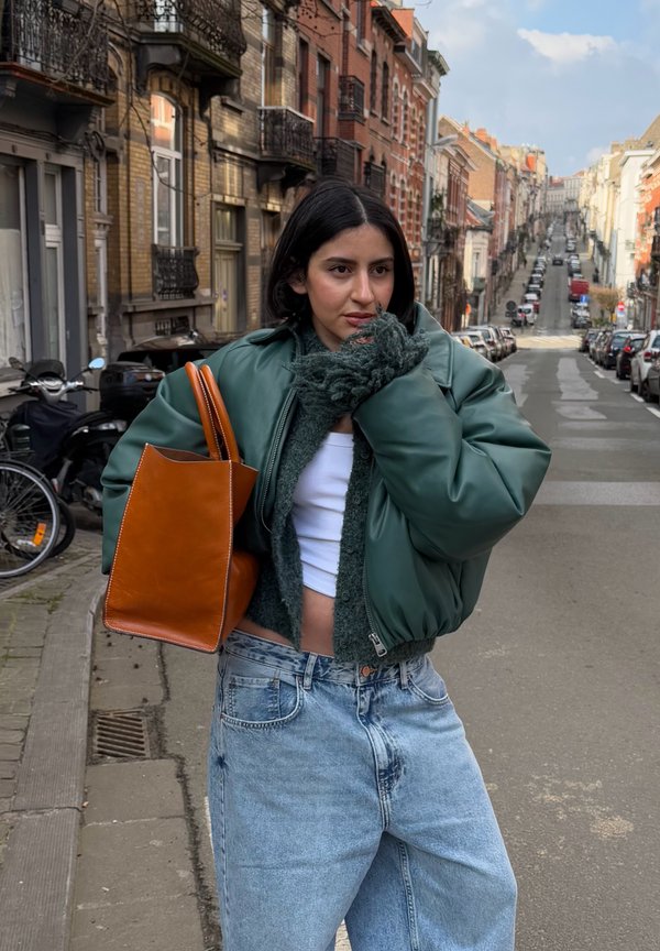 Giubbotto bomber in pelle verde, borsa a tracolla in pelle marrone, top corto bianco, maglione verde peloso, jeans in denim blu chiaro, sfondo urbano.