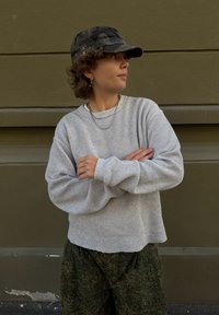 Grau strukturierter Pullover mit oversized Passform, kombiniert mit grünen Camouflage-Hosen. Eine dunkle Camouflage-Mütze rundet das Outfit ab. Schlichte silberne Kettenaccessoires.