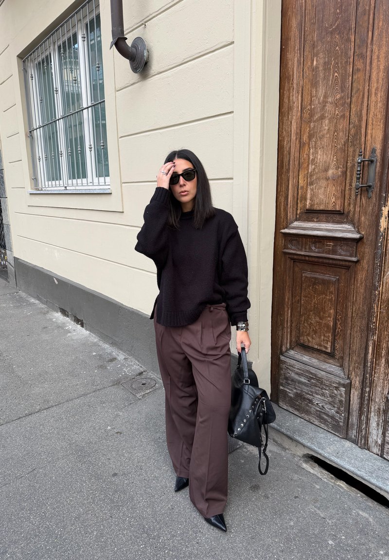 Mulher usando óculos de sol, suéter preto, calças largas castanhas e sapatos pretos pontiagudos, segurando uma mala preta, em pé junto a uma porta de madeira numa rua.